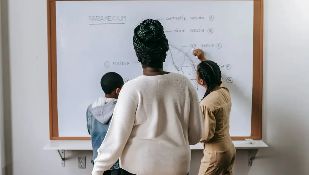 Professora escrevendo na lousa enquanto alunos acompanham a explicação
